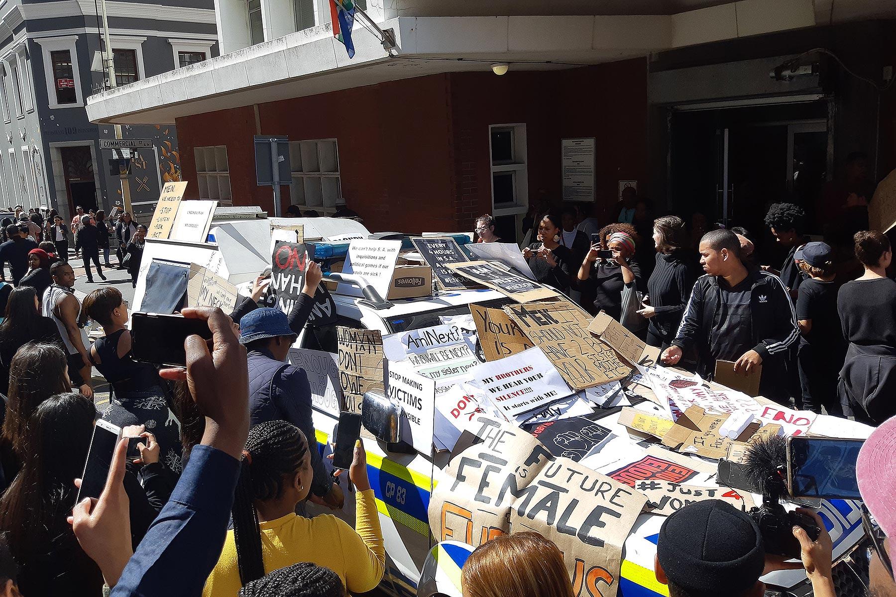In Kapstadt (Südafrika) wird nach dem Tod von Uyinene Mrwetyana gegen Frauenmorde protestiert. Dabei bedecken Demonstrierende ein Polizeiauto mit Protestschildern. Foto: Discott, via Wikimedia Commons (CC-BY-SA)
